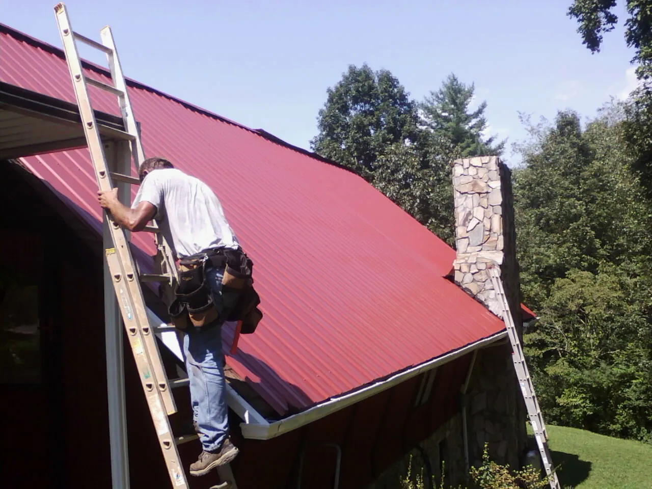 Roofer on a ladder beside a red metal roof and stone chimney — Soffit & Fascia Repair by Roofing Brothers in Gate